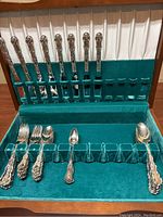 Whole silverware set including knives, forks, and spoons in green velvet-lined case showing ornate handles and arrangement.