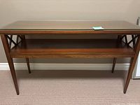 Front view of the wooden sofa table with glass top and lower tier shelf, showing the decorative X braces on the side panels and the tapered wooden legs.