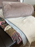 Photo showing three folded throw blankets: taupe plush blanket on top, beige and tapestry style blanket with fringe beneath.