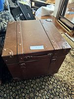 Single chestnut colored storage trunk with metal corner reinforcements and handles shown from the top and sides, placed on a patterned carpet.