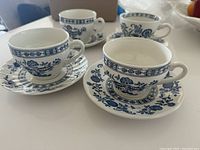 Four blue onion porcelain teacups with their matching saucers displayed on a white surface. The cups and saucers feature classic blue floral and onion botanical patterns on white porcelain.