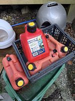 Four red gasoline canisters grouped in a black plastic crate; three older faded ones and one newer one with label.
