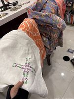 Four different tablecloths draped over two chairs: white embroidered linen, large orange cotton, blue floral, and orange-pink striped cloth.
