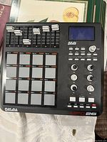 Top view of AKAI Professional MPD26 MIDI controller showing 16 large grey pads, 6 sliders, 6 knobs, LCD screen and function buttons.