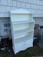 Front and angled views showing three open shelves and bottom cupboard with two doors, painted white with beadboard back paneling and decorative top finish.
