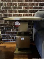 Side view of the table showing brass base and quartz top, with Choix Décoratifs tag hanging and visible brick background.