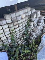 Stacks of square concrete paving bricks piled outdoors with some ivy growing nearby, showing weather exposure and moss.
