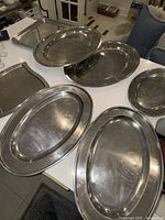 Seven stainless steel catering trays displayed on a white table showing varying shapes and sizes including oval, round and rectangular trays.