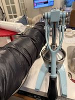 Close-up photo of the light blue metal citrus squeezer with black handle on a table, showing the juicing cone and front pressing arm.