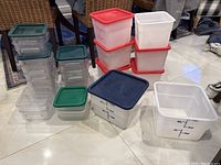Photo showing multiple clear plastic food storage containers with green, red, and blue lids on a tiled floor. Containers have volume markings and vary in size.