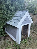 Photo showing side angle of large gray wooden dog house with white trim and shingled roof next to bushes on grass.