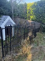Black metal garden gate and attached fence panels outdoors, partially overgrown with grass and plants.