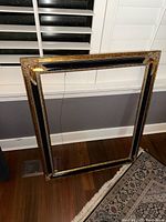 Large rectangular decorative frame with gold ornamental corners and black accents, leaning against a wall near a patterned rug and white shutters.