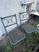 Two green metal folding garden chairs placed outside against a wall, showing seats and backs with mesh and circular decoration.