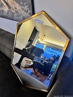 Photo of an octagonal wall mirror with a gold-tone frame placed leaning against a wall reflecting a room with furniture and windows.