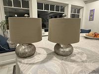 Photo shows a pair of table lamps with metallic textured sea urchin style bases and beige fabric shades on a table indoors.