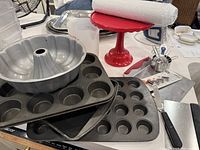 Photo showing the new red cake stand, two black non-stick muffin pans, silver bundt cake pan, plastic measuring cup, metal sifter, set of cake decorating tips, pastry knife, and metal mesh roll on the table.