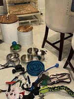 Photo showing four stainless steel dog bowls of different sizes, two white food storage containers with wooden lids, various dog toys including balls and chew toys, three nail clippers, grooming brushes/combs, and several leashes arranged on the floor in a dining room area.