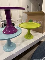 Three 9-inch round cake stands stacked and displayed on a counter, purple on top, lime green in middle, light blue at bottom, original retail box visible.