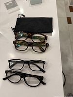 Four pairs of reading glasses on a white surface, two tortoiseshell frames, and two black frames, with a black soft pouch above.