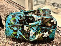 Roxy duffle bag lying flat on a patterned carpet, showing the floral design and handles with Velcro closure.