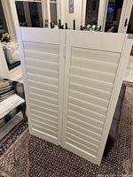 Two white wood shutter panels connected by hinges with louvers, leaning on a couch in a living room setting.