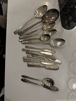 Photo showing an assortment of silver plated cutlery including large spoons, small spoons, multiple butter knives, and a slotted spoon, all with varying handle patterns and some tarnishing.