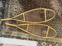 Photo showing the pair of full size wooden snowshoes on a patterned carpet, revealing the traditional design with rawhide webbing and smooth wooden frames in new condition.