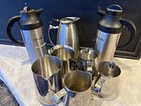 Photo showing 3 thermal carafes labeled 'CREAM' and 5 stainless steel jugs on fabric surface.