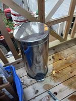Closed view of a tall, cylindrical stainless steel trash can with foot pedal and reflective surface, placed on a wooden deck outdoors.