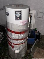 Daylight photo of white Bradford White Defender Safety System water heater tank with red tape securing plastic insulation, placed outdoors next to a wall and blue container.