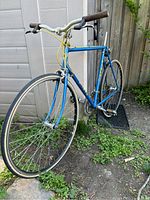 Full side angled view of blue vintage Miele road bicycle with drop handlebars, yellow cables and narrow tires placed outdoors on an uneven grassy and paved surface near a fence.