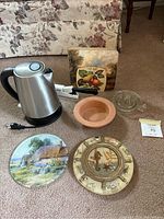 Photo showing all items in the lot including electric kettle, ceramic wine bottle dish, glass juicer, cutting tool, and trivets.