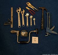 Photo shows assortment of hand tools laid out on a blue carpet including combination square, wrenches, locking pliers, hammer, caulking gun, tape measure, and hand drill.
