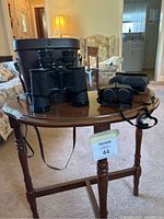 Two pairs of binoculars on a wooden side table: larger Bell & Howell pair with case and smaller Bushnell pair with strap.
