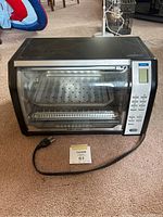 Full view of the Black & Decker convection oven showing black body, glass door, visible trays inside, power cord placed outside, and original user manual underneath.