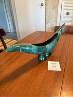 Side angled view of long, green swirled pottery lamp vase with small pedestal base on wooden table.