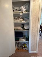 Closet with shelves holding folded linens including tablecloths, towels, sheets and pillowcases, mattress cover, mostly white and neutral colors.