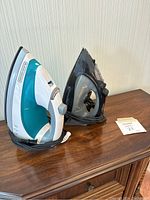 Two irons side by side on a wooden surface, one Black & Decker primarily white and teal, one black Proctor Silex, showing overall condition and appearance.