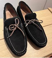 Front angle view of the pair of black suede UGG loafers with brown leather ties and white stitching details displayed on a wooden surface.