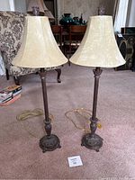Two matching floor lamps standing on carpet with beige bell-shaped fabric shades, dark metal bases with ornamental detailing.