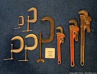Overview of three pipe wrenches with red handles next to five metal C clamps of varied sizes and vintage on blue fabric background
