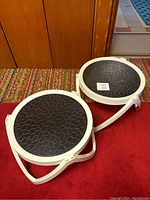 Two white plastic side tables with black removable round tops, placed on a multicolor rug and red carpet, in front of a wooden paneled wall.
