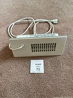 White rectangular floor register booster fan heater with vent slats and power cord attached lying on carpeted floor.