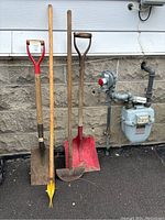 Four garden tools standing upright against a stone wall: two shovels, one red metal shovel, one yellow edging tool, and one dandelion remover.