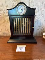 Closed black wooden tabletop clock with curved top, round clock face with Roman numerals, and glass front door with vertical brass bars behind it, resting on wooden surface.
