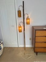 Full view of the vintage floor-to-ceiling lamp showing three lit amber glass shades and the tall dark wood pole resting against the wall.
