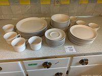Set of white ceramic dishes arranged on countertop showing all items including saucers, mugs, side plates, bowls, serving bowl, and dinner plates.