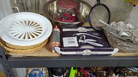 Photo of shelf with woven basket paper plate holder, clear glass pitcher, metal mixing bowl, metal cookie press, boxed corkscrew, and clear glass and crystal bowls with utensils inside.