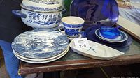 Photo showing a covered pot with blue scenic design, blue and white dinner plates, a mug, and a small boat-shaped dish